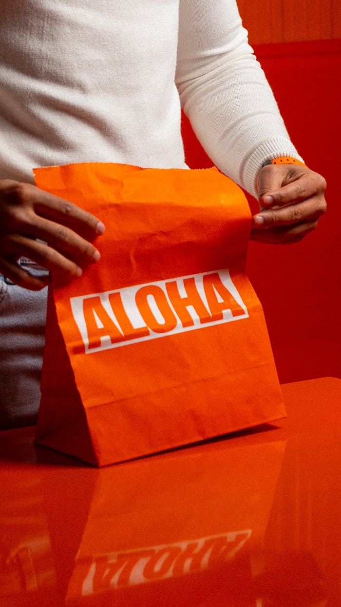 The Art of Drawing Readers In: Your attractive post title goes here Close-up of hands holding a bright orange 'ALOHA' paper bag on an orange surface. Indoor setting.